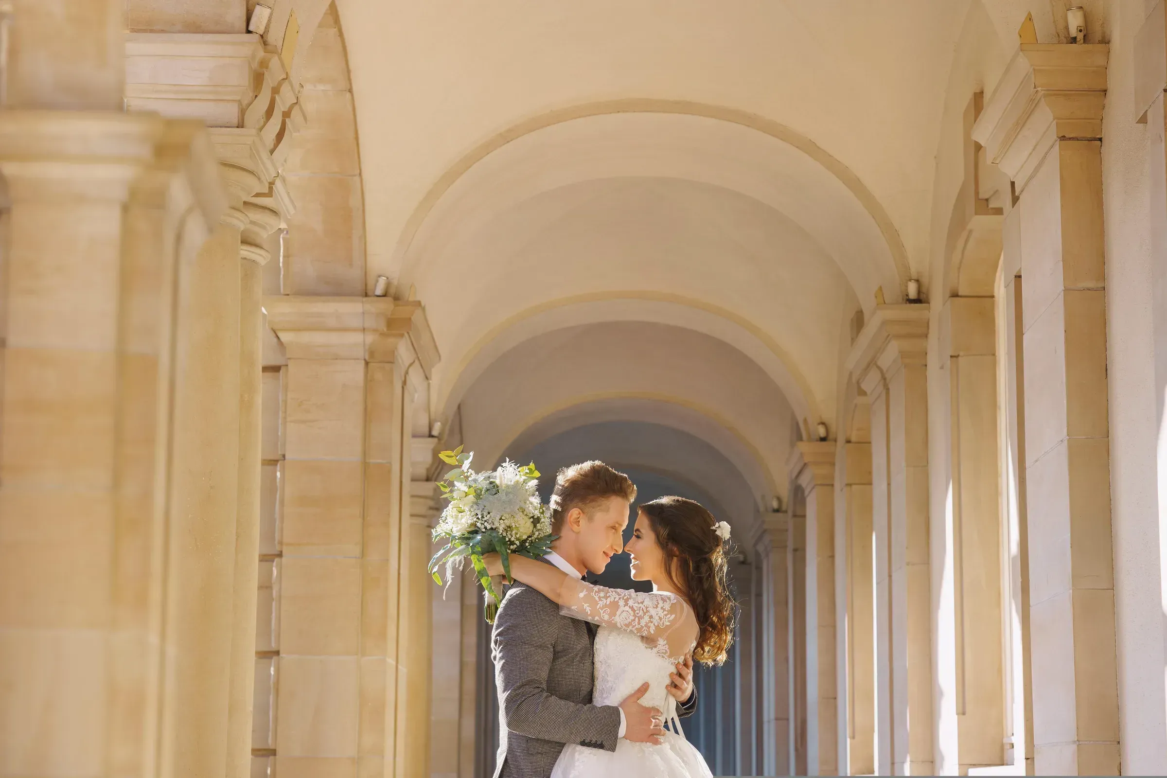 A bride and groom holding each other under a long stone colonnade.