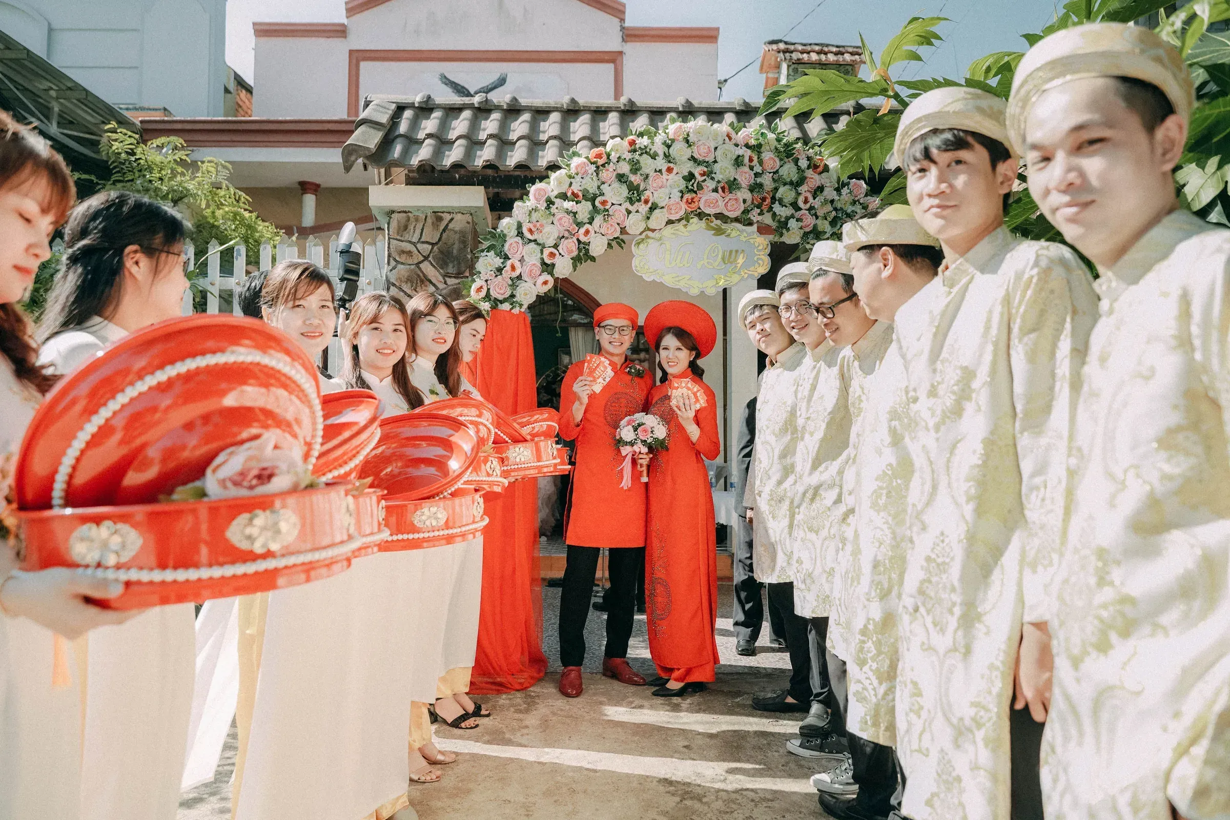 A bride and groom in red under a flower arch, with guests lined up.